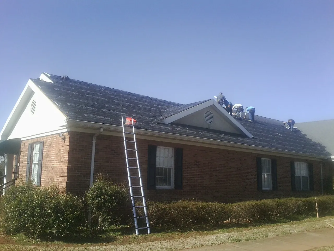Crew tearing shingles off a brick ranch roof with a ladder set — Emergency Roof Repair work by our crew in New Hampton